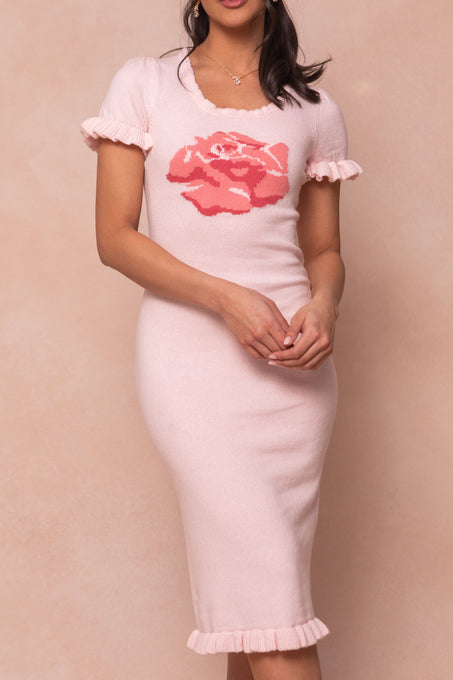 A woman wears the Ivy City Co Rosie Knit Dress, a maternity-friendly pale pink dress with ruffled sleeves, a fitted silhouette, and a bold pink rose graphic on the chest, standing against a soft beige background.
