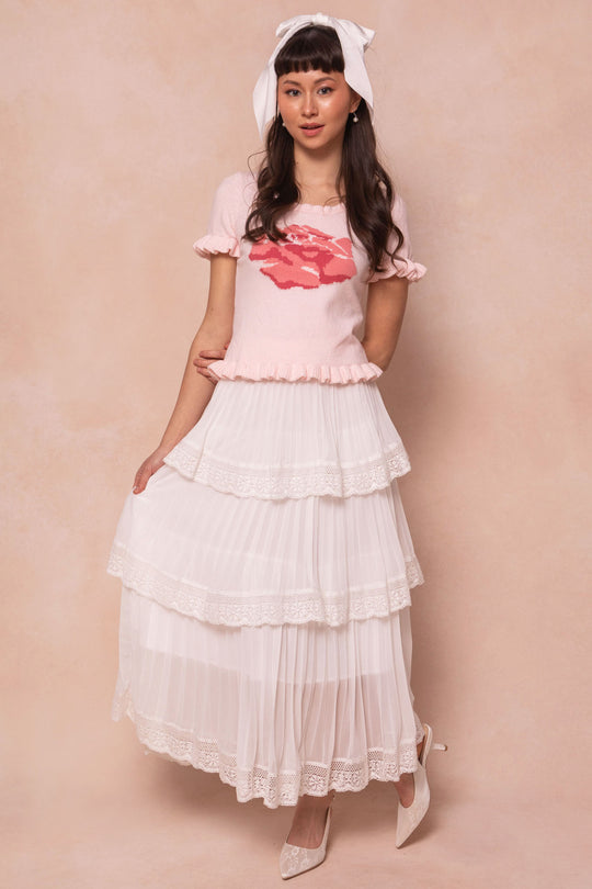 A woman poses against a soft beige background wearing the Rosie Knit Top by Ivy City Co in pink with a red graphic, paired with a white tiered lace midi skirt with ruffles, white heels, and a large white headband.