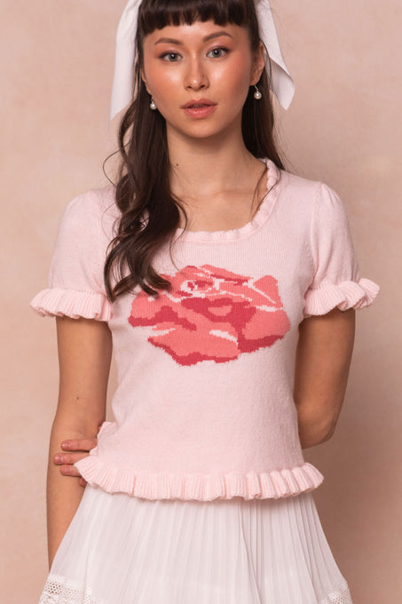 A woman with long brown hair and bangs wears the Ivy City Co Rosie Knit Top, featuring ruffle detail and a large rose design, paired with a white pleated skirt. She completes her look with pearl earrings and a white hair ribbon.