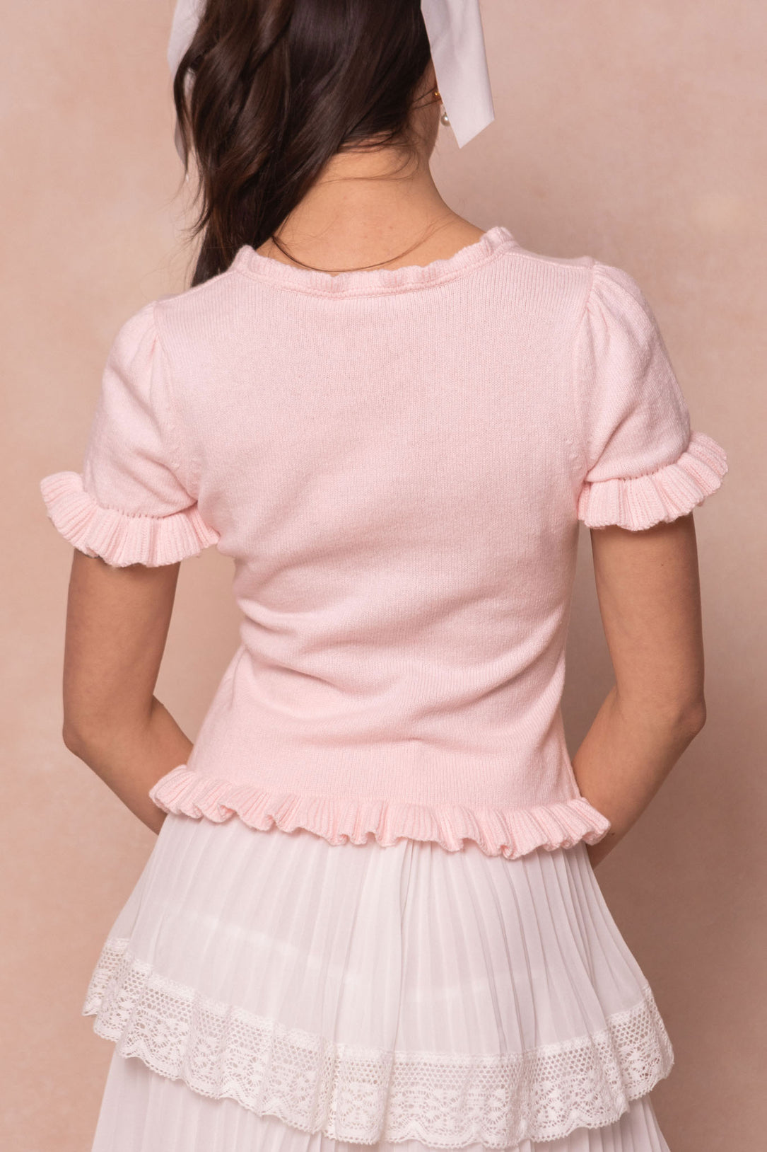 A woman faces away, wearing the Ivy City Co Rosie Knit Top in pink with ruffled edges and a white pleated skirt with lace trim. Her brown hair is tied with a white ribbon, set against a soft beige background.
