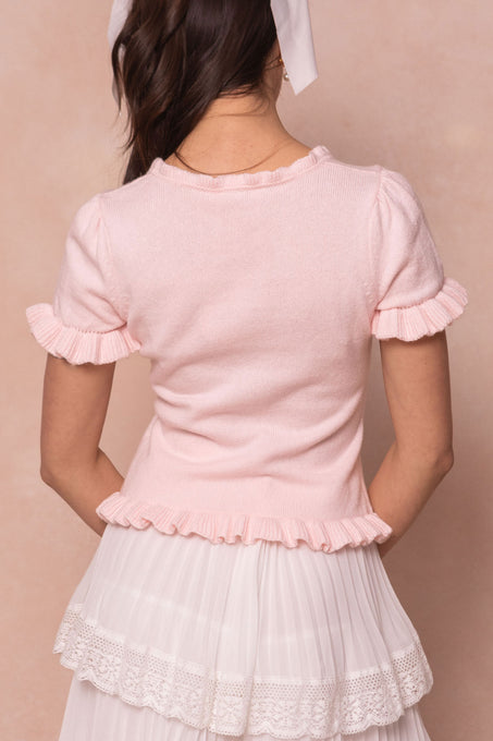 A woman faces away, wearing the Ivy City Co Rosie Knit Top in pink with ruffled edges and a white pleated skirt with lace trim. Her brown hair is tied with a white ribbon, set against a soft beige background.