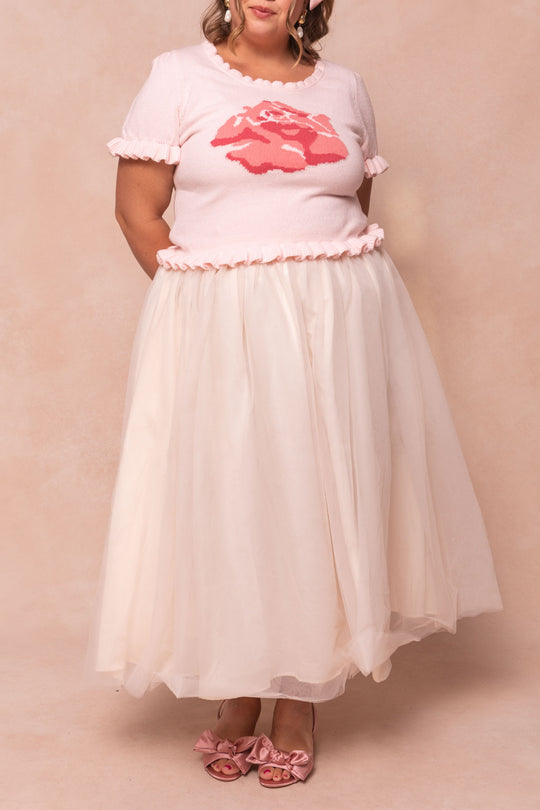 A person wears the Ivy City Co Rosie Knit Top—a pink knit top with a red rose pattern and ruffled collar and sleeves—paired with a voluminous light pink tulle skirt and bow sandals, standing against a soft beige background.