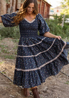 A woman stands outdoors among rustic buildings and greenery, gently holding out the skirt of the Roselyn Dress in Midnight Lovelorn Ditsy by Ivy City Co. This blue, floral, long-sleeve cotton midi dress features a graceful silhouette and is also maternity friendly. She gazes down at her flowing skirt, embodying effortless elegance.