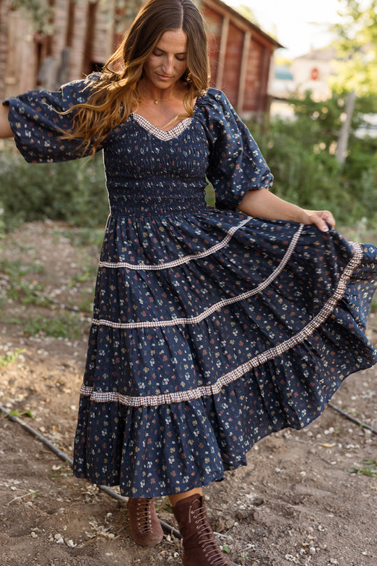 A woman stands outdoors among rustic buildings and greenery, gently holding out the skirt of the Roselyn Dress in Midnight Lovelorn Ditsy by Ivy City Co. This blue, floral, long-sleeve cotton midi dress features a graceful silhouette and is also maternity friendly. She gazes down at her flowing skirt, embodying effortless elegance.