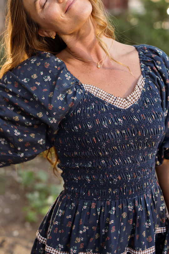 A woman wearing the Ivy City Co Roselyn Dress in Midnight Lovelorn Ditsy—a maternity-friendly, navy floral cotton midi dress featuring puff sleeves, a smocked bodice, and gingham trim—smiles with closed eyes outdoors in sunlight.