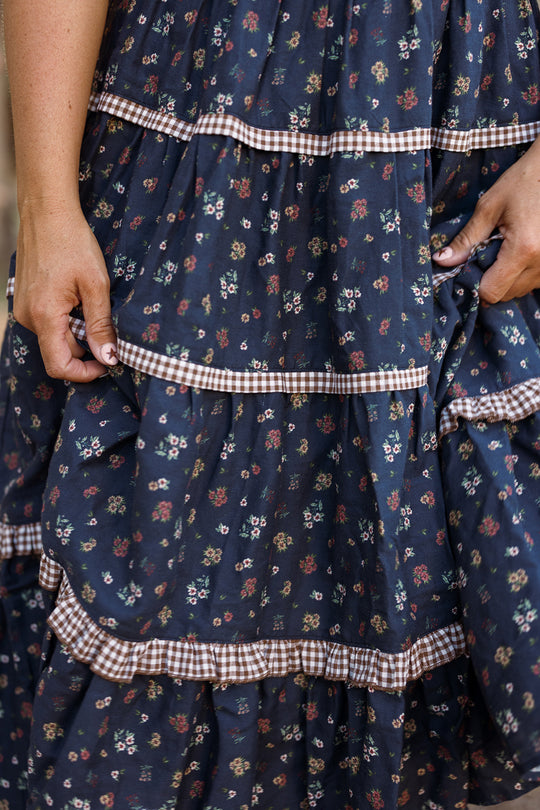 A close-up of a person’s hands holding the Ivy City Co Roselyn Dress in Midnight Lovelorn Ditsy—a navy blue cotton midi dress adorned with small floral prints and gingham trim. This maternity-friendly dress features layered ruffles, with the person’s arms partially visible.