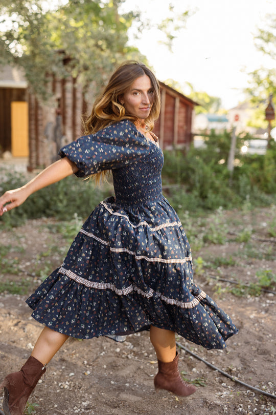 A woman twirls outdoors near rustic buildings, surrounded by greenery and sunlight, wearing the Ivy City Co Roselyn Dress in Midnight Lovelorn Ditsy—a blue patterned cotton midi dress that is maternity friendly.