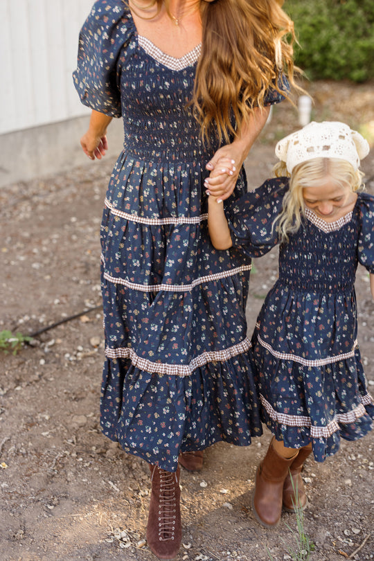 A woman and a young girl hold hands while walking outdoors, both wearing matching Ivy City Co Roselyn Dresses in Midnight Lovelorn Ditsy—a navy blue floral print. The girl pairs her cotton, maternity-friendly midi dress with a white headscarf, and both wear brown boots. Their faces are not visible.