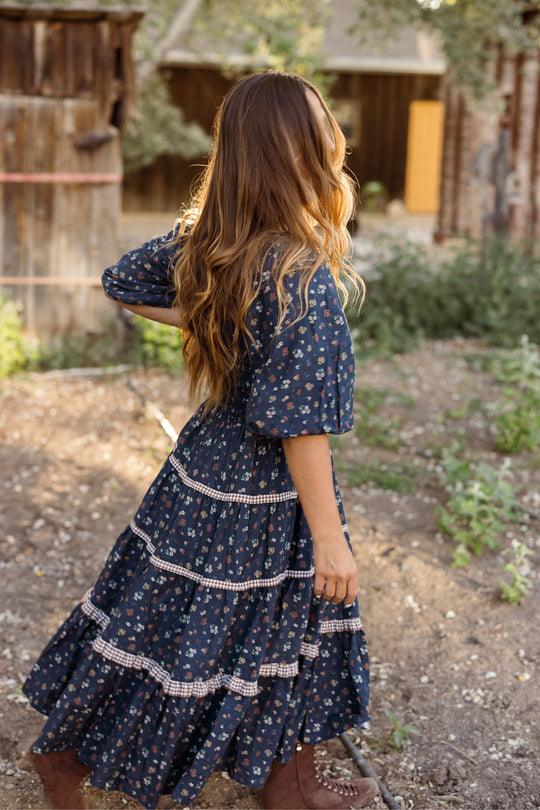A woman with long brown hair, wearing the Ivy City Co Roselyn Dress in Midnight Lovelorn Ditsy, walks outdoors on a dirt path facing away. She is surrounded by plants, with rustic wooden buildings and trees in the background. This blue floral cotton midi dress is both stylish and maternity friendly.