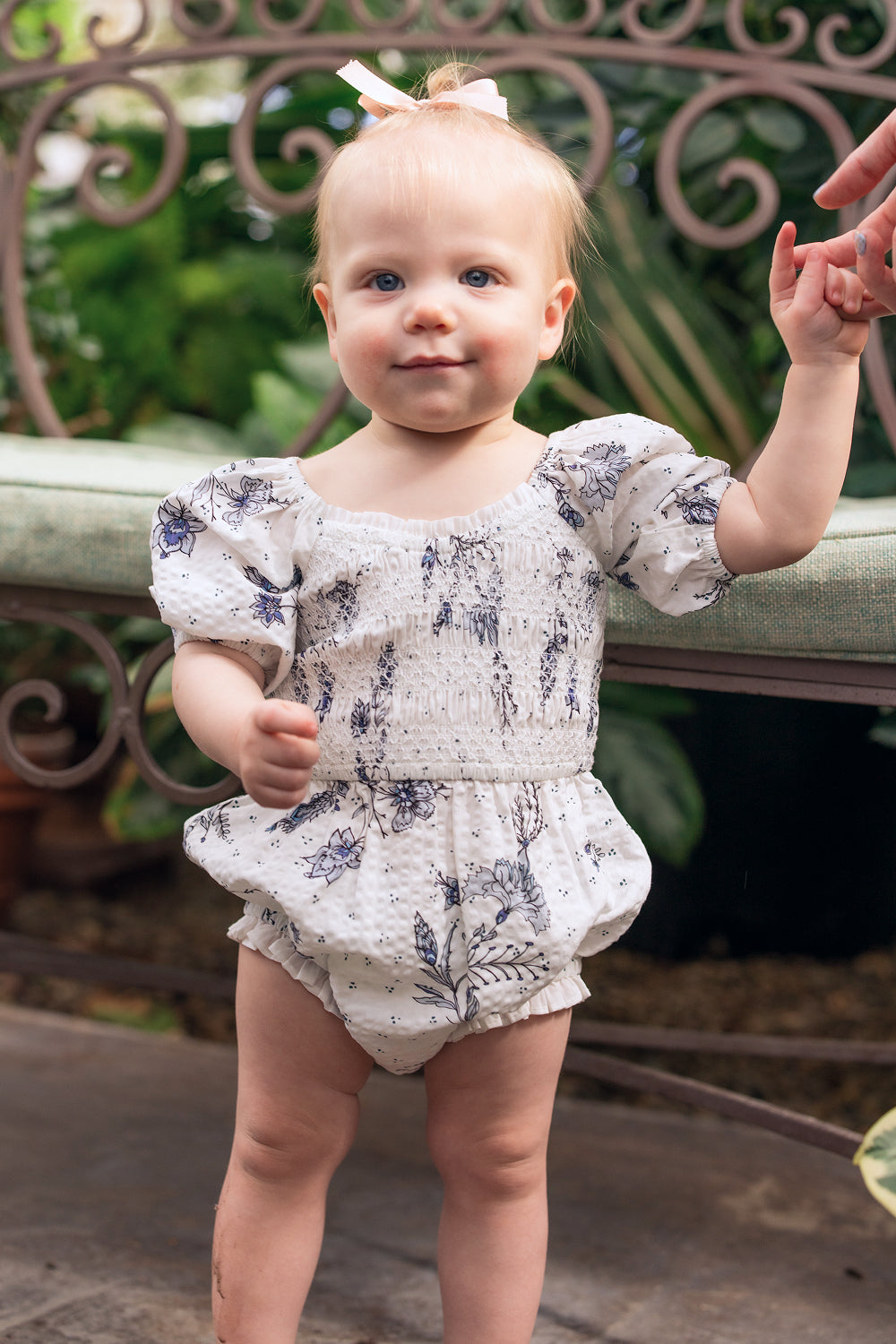 A smiling baby girl with blue eyes and a pink bow stands outdoors in the Ivy City Co Baby Roselyn Romper in Blue Enchanted Floral, holding an adult’s hand by an iron bench and greenery—perfect for precious Mom & Me moments.