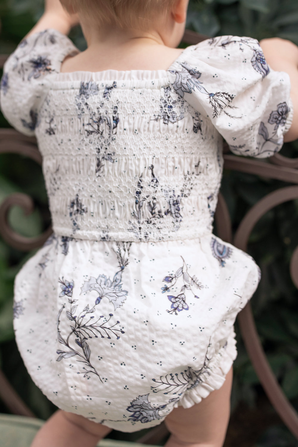 A baby in the Ivy City Co Baby Roselyn Romper in Blue Enchanted Floral with puffed sleeves stands by a metal railing amid greenery—perfect for matching Mom & Me moments.
