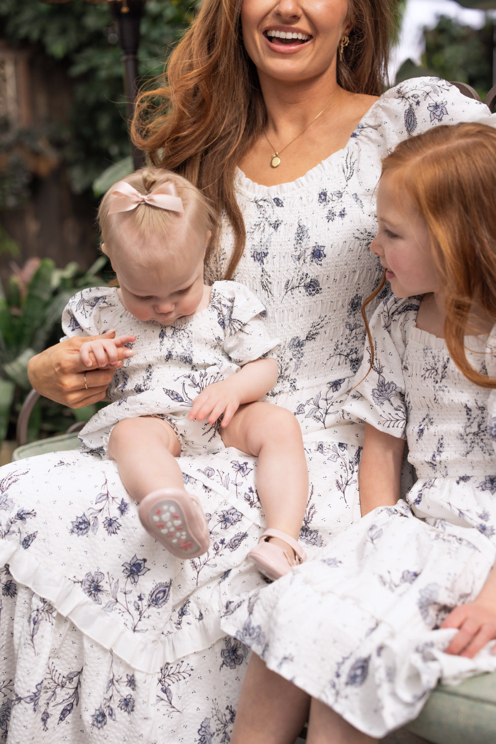 A woman, baby, and young girl sit together joyfully in matching blue enchanted floral dresses. The baby wears a bow and the adorable Ivy City Co Baby Roselyn Romper in Blue Enchanted Floral completes their sweet Mom & Me ensemble.