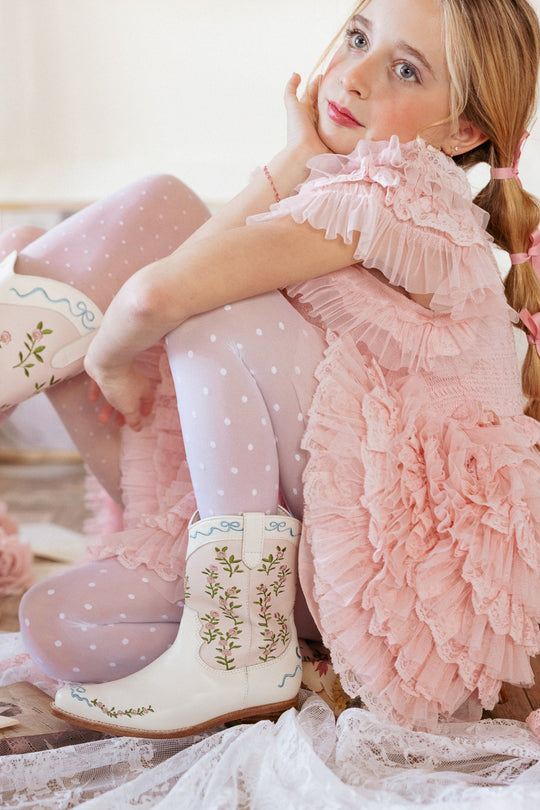 A young girl in a ruffled pink dress and light pink polka dot tights sits thoughtfully, her look finished with Ivy City Cos Mini Rosemary Cowgirl Boots, featuring an embroidered floral pattern.