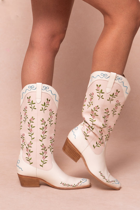 A person wearing Ivy City Cos Rosemary Cowgirl Boots—white with embroidered florals, light blue trim, and mid-height wooden heels—stands against a pale background.