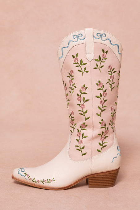 The Rosemary Cowgirl Boots by Ivy City Co are light pink with embroidered green stems, pink flowers, blue trim at the top and toe, and a low wooden heel, shown on a pale pink background.