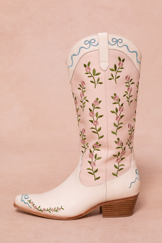 The Rosemary Cowgirl Boots by Ivy City Co are light pink with embroidered green stems, pink flowers, blue trim at the top and toe, and a low wooden heel, shown on a pale pink background.