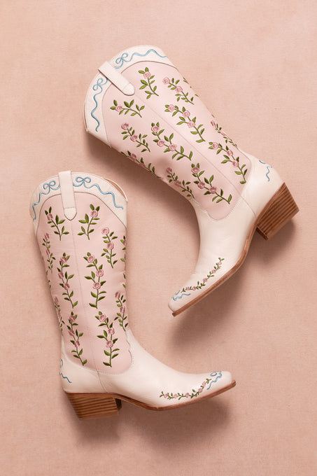 The Rosemary Cowgirl Boots by Ivy City Co are light pink leather boots with an embroidered floral pattern and blue trim, displayed on a soft pink background, both boots lying on their sides with heels angled outward.