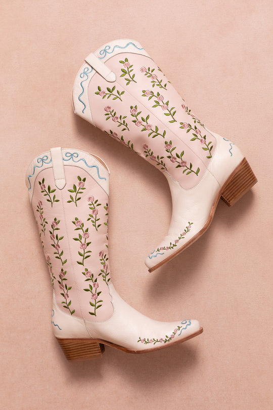 The Rosemary Cowgirl Boots by Ivy City Co are light pink leather boots with an embroidered floral pattern and blue trim, displayed on a soft pink background, both boots lying on their sides with heels angled outward.