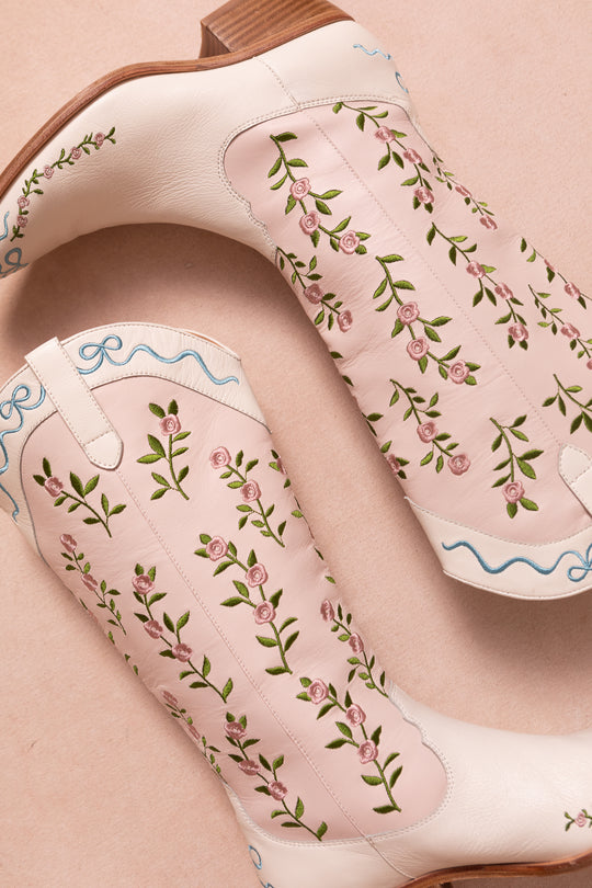 The Rosemary Cowgirl Boots by Ivy City Co feature pink leather with light brown heels, embroidered green vines and small pink flowers, blue swirl accents near the top, and are displayed on a soft pink surface.