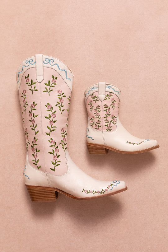 The Mini Rosemary Cowgirl Boots by Ivy City Co feature wooden heels and an embroidered floral design with green vines, pink flowers, and blue ribbons on light pink. Shown on a soft pink background.