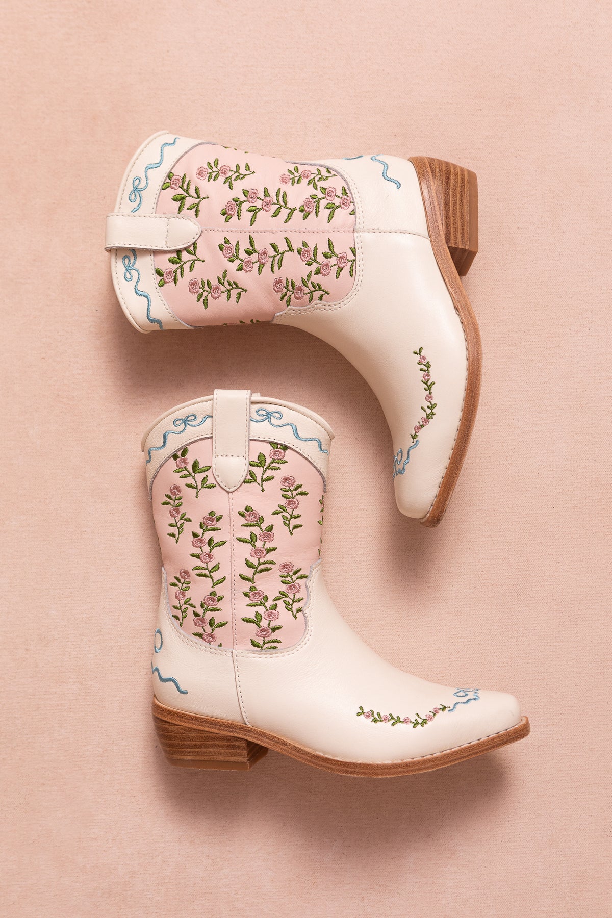 Two Mini Rosemary Cowgirl Boots in Cream & Pink by Ivy City Co, featuring wooden heels, floral embroidery, and delicate pink trim, are placed on a soft pink background.