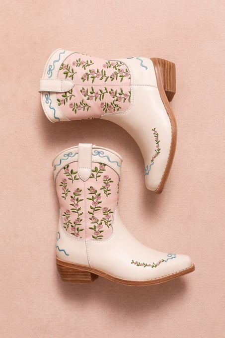 The Mini Rosemary Cowgirl Boots by Ivy City Co in cream feature wooden heels, pink panels, and blue swirled floral embroidery, shown on a light pink background.
