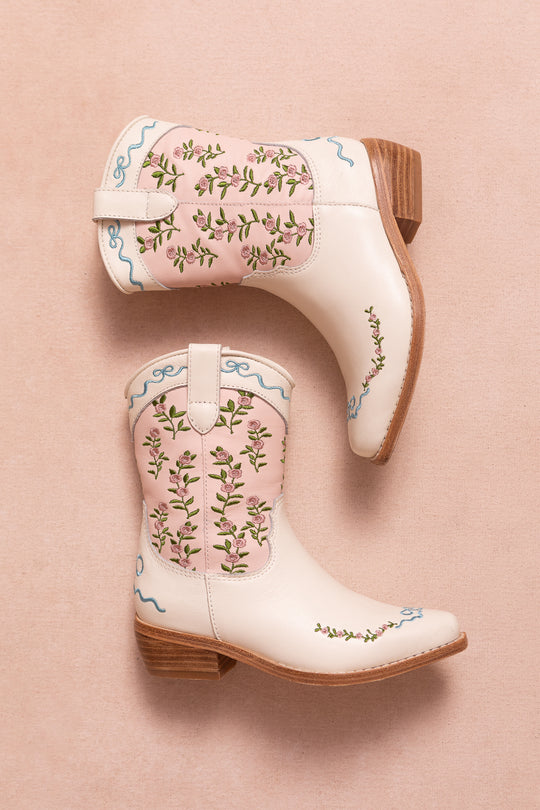 The Mini Rosemary Cowgirl Boots by Ivy City Co in cream feature wooden heels, pink panels, and blue swirled floral embroidery, shown on a light pink background.