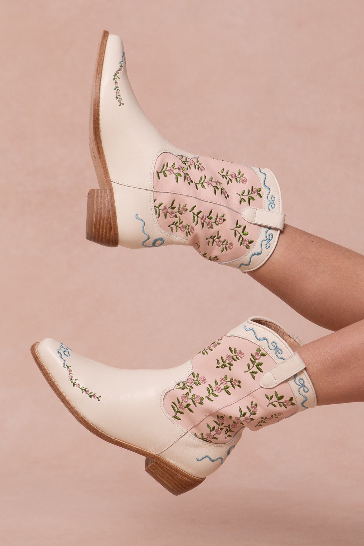 A pair of cream and pink cowgirl boots with floral embroidery, pink trim, and wooden heels, modeled by a person lying down with legs raised against a soft pink background.