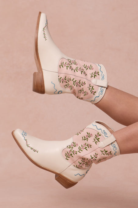 A person wearing Ivy City Co’s Mini Rosemary Cowgirl Boots in cream and light pink with floral embroidery and blue stitching, legs raised against a blush pink background.