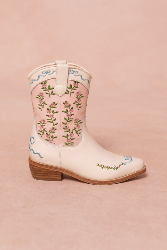 The Ivy City Co Mini Rosemary Cowgirl Boots showcase a light pink and cream design, floral embroidery, blue stitching, a brown low heel, and side pull tab, all set against a soft pink background.