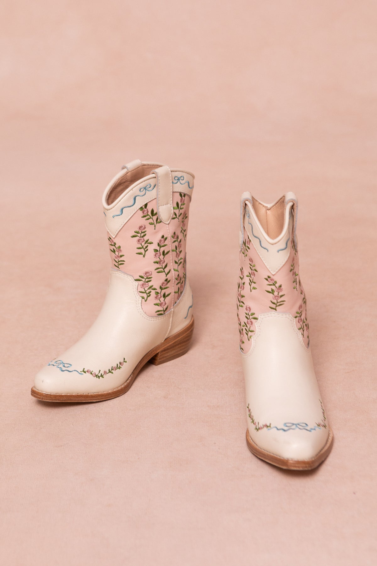 A pair of white leather cowgirl boots with cream and pink floral embroidery, blue decorative accents, and delicate pink trim, displayed on a soft beige surface.