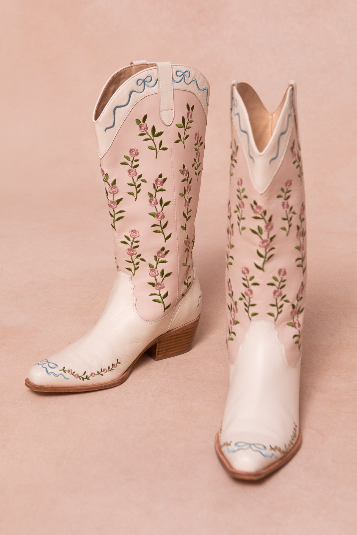 The Rosemary Cowgirl Boots by Ivy City Co are cream-colored with light brown heels, featuring pink floral embroidery, green vines, and blue ribbons on the tops and toes set against a neutral background.