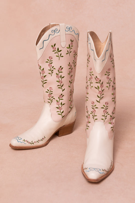 The Rosemary Cowgirl Boots by Ivy City Co are cream-colored with light brown heels, featuring pink floral embroidery, green vines, and blue ribbons on the tops and toes set against a neutral background.