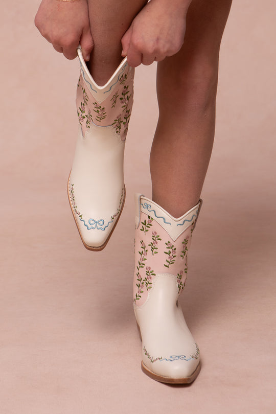 Against a soft beige background, only hands and legs are visible as someone slips on Ivy City Cos Mini Rosemary Cowgirl Boots—white leather boots featuring embroidered florals and light blue bows.