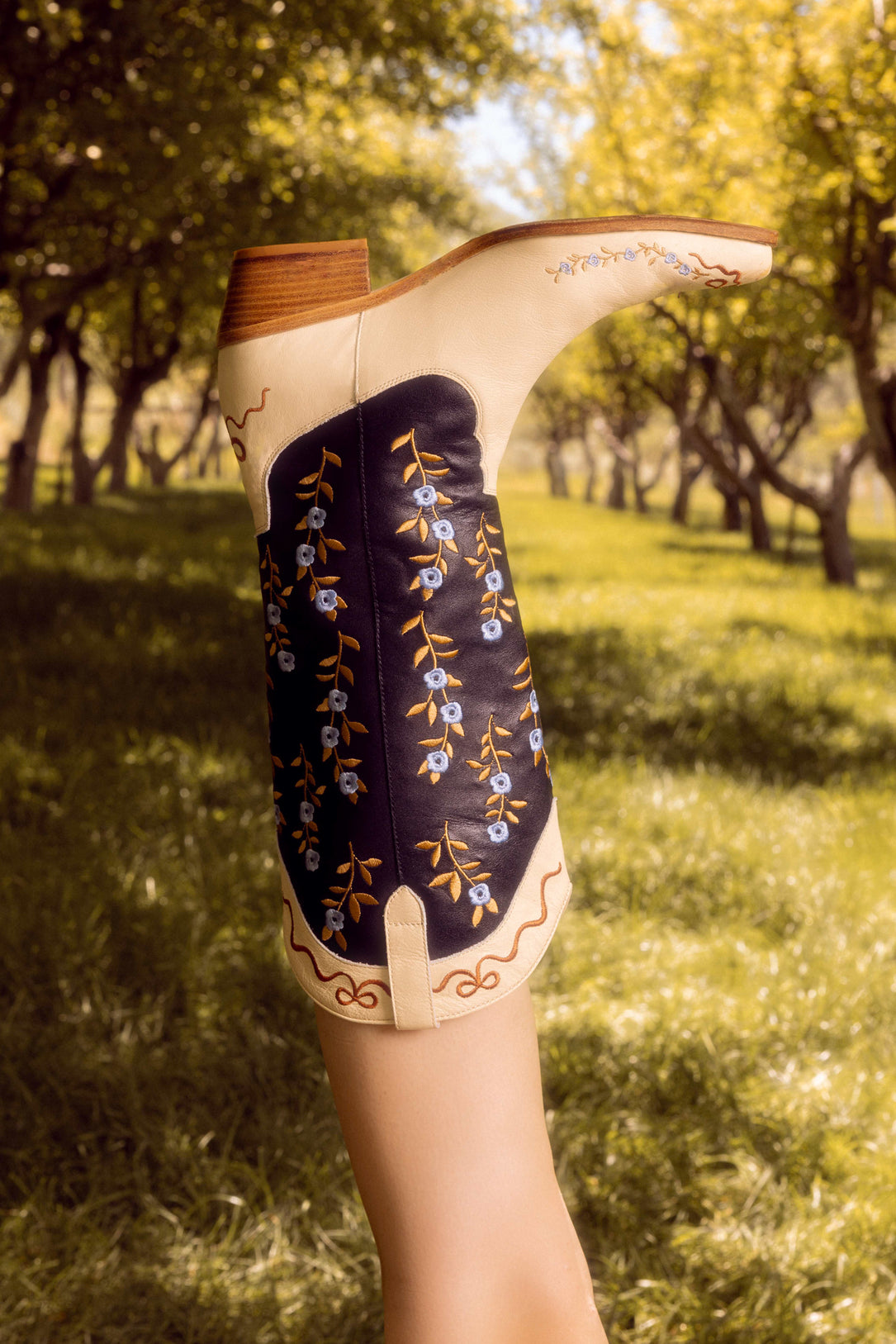 A person raises their leg outdoors, showcasing Ivy City Co’s Rosemary Cowgirl Boots in Cream & Navy with blue and yellow floral embroidery, set against a backdrop of trees and sunlight.