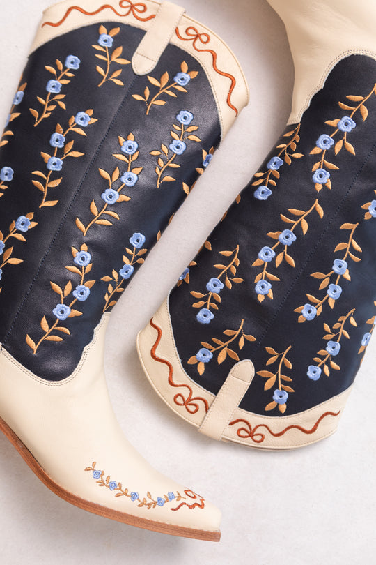 Close-up of Ivy City Cos Rosemary Cowgirl Boots in Cream & Navy, featuring blue and yellow floral embroidery, black accents, and brown decorative stitching along the edges.