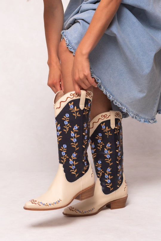 A person in a light blue, frayed-edge dress adjusts Ivy City Cos Rosemary Cowgirl Boots in Cream & Navy, featuring blue floral embroidery and tan soles, against a light background.