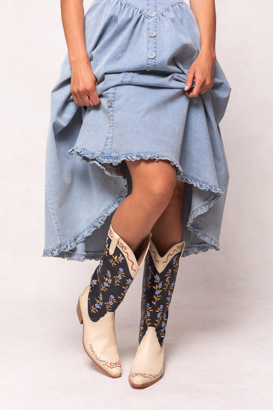 A person stands against a plain light background wearing a light blue high-low denim skirt and Ivy City Co’s Rosemary Cowgirl Boots in Cream & Navy.