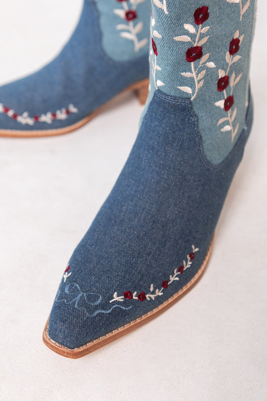 Close-up of Ivy City Cos Rosemary Cowgirl Boots in Denim, showcasing embroidered red flowers and white leaves on the shaft and toe, with a light brown sole and detailed stitching.