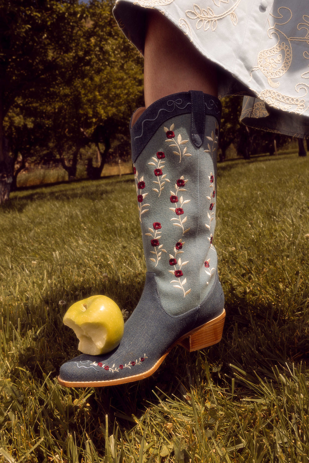 A person wears Ivy City Cos Rosemary Cowgirl Boots in Denim, featuring blue floral embroidery, standing on grass beside a bitten green apple. The edge of a light blue embroidered skirt is also visible.