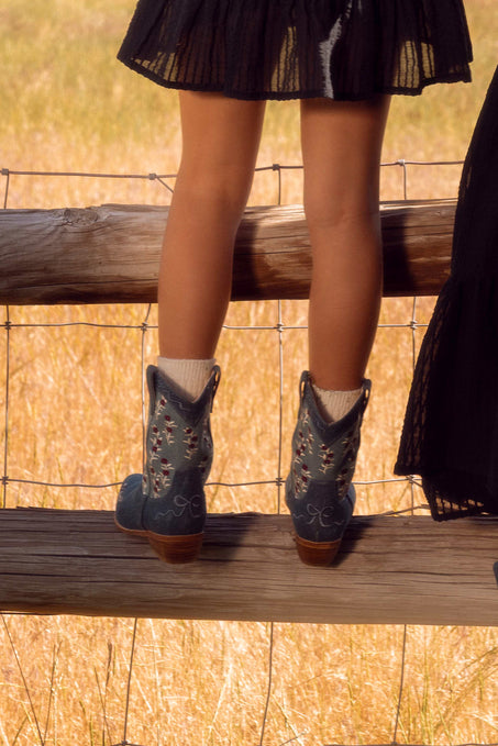 A child wearing a black skirt, white socks, and Ivy City Co’s Mini Rosemary Cowgirl Boots in Denim stands on a wooden fence, facing away, with dry grass fields in the background.