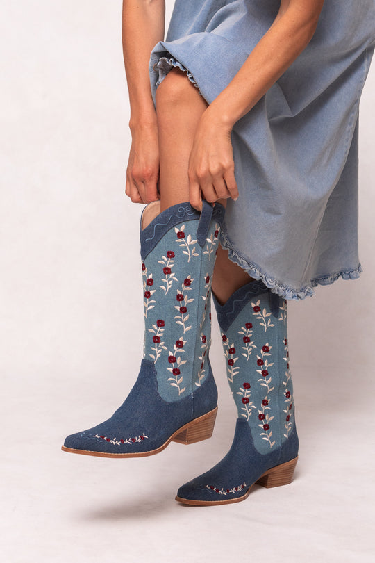 A person in a light blue dress adjusts Ivy City Cos Rosemary Cowgirl Boots in Denim, which feature red and white floral embroidery, a pointed toe, and a low wooden heel.