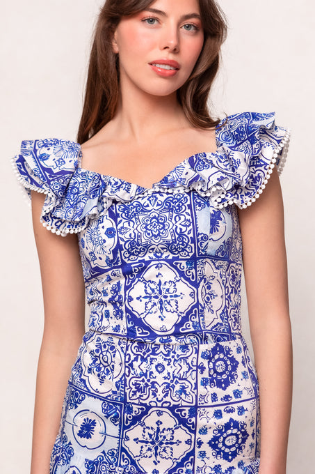A woman stands against a plain background wearing the Ivy City Co Rosina Blouse in Blue Tile—a cotton top with flutter sleeves, tile-inspired blue and white patterns, and white pompom trim along the ruffles.