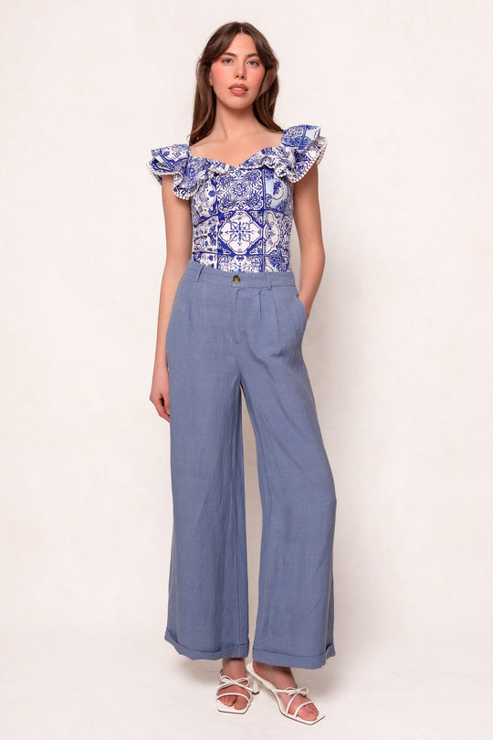 A woman poses with her hands in her pockets, wearing the Ivy City Co Rosina Blouse in Blue Tile, flutter sleeves, high-waisted light blue wide-leg pants, and white strappy sandals against a plain background.
