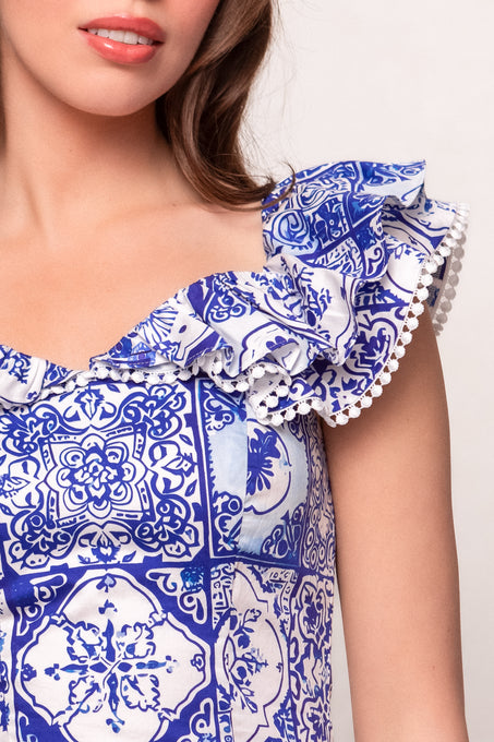 A woman wears the Ivy City Co Rosina Blouse in Blue Tile, featuring intricate blue and white tile patterns, ruffled flutter sleeves, and small white pom-poms. Only her lower face, neck, and upper torso are visible.