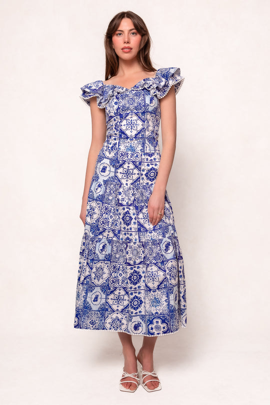 A woman wearing the Ivy City Co Rosina Blouse in Blue Tile stands facing forward against a plain light background. She has long hair and wears white sandals.