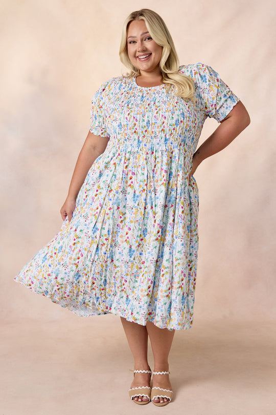 A smiling woman with long blonde hair stands against a neutral background, holding out the skirt of the Ivy City Co Amelie Dress in Floral Fields—a light blue, floral midi dress. She completes the look with white sandals.