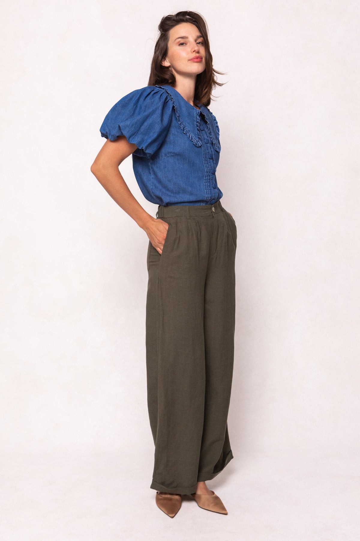 A woman wears high-waisted olive green Veronica Pants in Green with a wide-leg fit, paired with a short-sleeved blue denim top with ruffled shoulders.