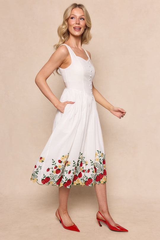 A woman with long blonde hair wears the Ivy City Co Clarissa Dress in Strawberry—a sleeveless white dress featuring colorful floral embroidery along the hem—paired with red pointed-toe heels, standing against a beige background, smiling and looking to the side.