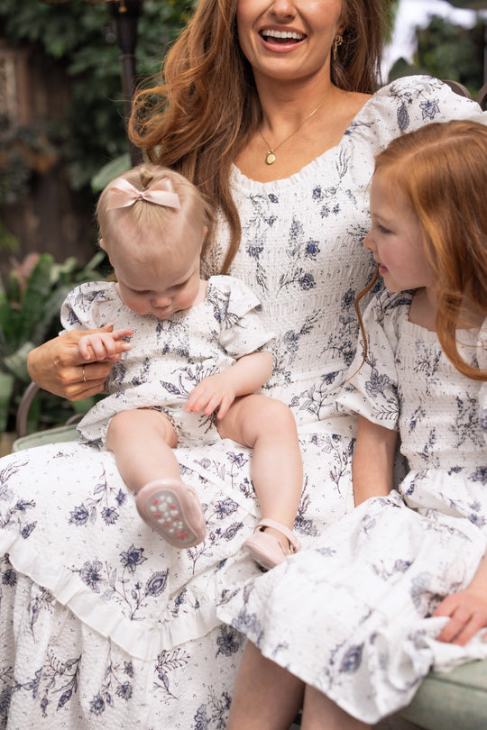 A woman sits outdoors holding two young girls in matching blue floral outfits. The baby wears the Ivy City Co Baby Roselyn Romper in Blue Enchanted Floral and looks down, while the older child, also dressed in a coordinating Ivy City Co Mom & Me set, gazes at her little sister. All three wear white and blue patterned ensembles from Ivy City Co, creating a charming family moment.